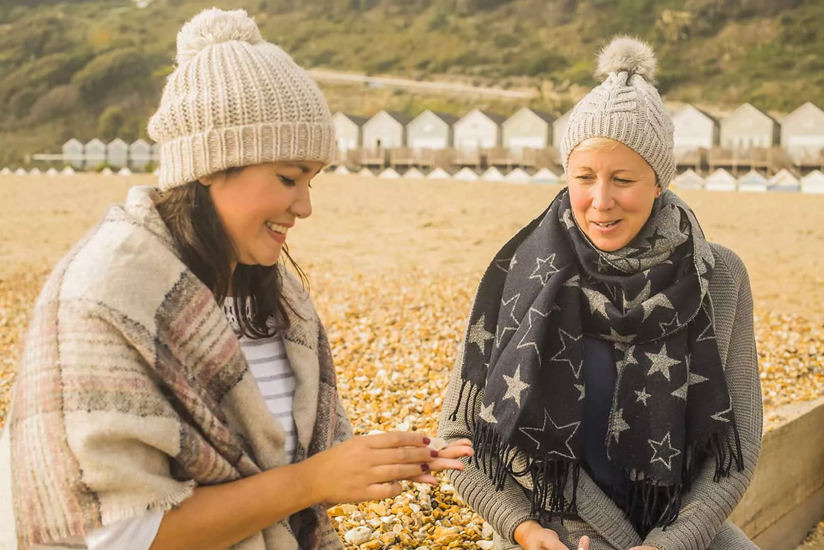 Two women sat on beach wrapped up warm with lodges in the background