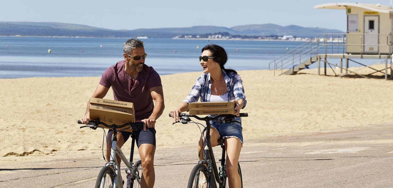 Two people riding bicycles along the prom at the beac carrying pizza boxes